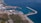 Aerial view of a coastal port with several ships docked. The area features a marina with numerous smaller boats, surrounded by industrial facilities. Further inland, green vegetation and urban buildings are visible. The coastline curves gently, bordered by clear blue waters.