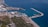 Aerial view of a coastal port with several ships docked. The area features a marina with numerous smaller boats, surrounded by industrial facilities. Further inland, green vegetation and urban buildings are visible. The coastline curves gently, bordered by clear blue waters.