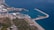 Aerial view of a coastal port with several ships docked. The area features a marina with numerous smaller boats, surrounded by industrial facilities. Further inland, green vegetation and urban buildings are visible. The coastline curves gently, bordered by clear blue waters.