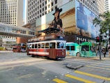 A vintage-style tram with a brown and cream color scheme is traveling through a busy urban street. The background features a high-rise building with a large advertisement showing a woman in a black hat. Other vehicles, including a green bus and a red double-decker bus, can also be seen alongside pedestrians.