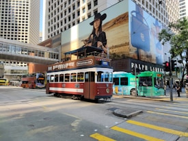 A vintage-style tram with a brown and cream color scheme is traveling through a busy urban street. The background features a high-rise building with a large advertisement showing a woman in a black hat. Other vehicles, including a green bus and a red double-decker bus, can also be seen alongside pedestrians.