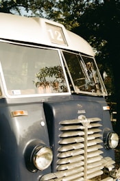 A vintage blue bus with a 'Mavag' logo is parked outdoors, featuring a large front grille, round headlights, and a sign displaying the number 12. Reflections of trees provide a natural backdrop.