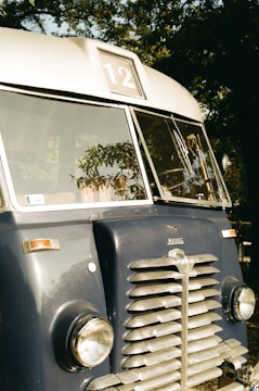 A vintage blue bus with a 'Mavag' logo is parked outdoors, featuring a large front grille, round headlights, and a sign displaying the number 12. Reflections of trees provide a natural backdrop.