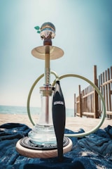 A hookah is placed on a beach setting with clear blue skies and the sea in the background. The glass base of the hookah is partially filled with water, and a hose is attached, draped around the setup. It is positioned on top of a blue cloth, with some wooden fencing seen to the right side.