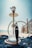 A hookah is placed on a beach setting with clear blue skies and the sea in the background. The glass base of the hookah is partially filled with water, and a hose is attached, draped around the setup. It is positioned on top of a blue cloth, with some wooden fencing seen to the right side.