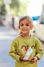A young girl with a cheerful expression, standing outdoors. She wears a green sweatshirt with a logo on it and holds a plastic container in her hands. The background is blurred, suggesting an urban setting.