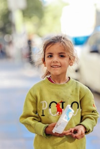 A young girl with a cheerful expression, standing outdoors. She wears a green sweatshirt with a logo on it and holds a plastic container in her hands. The background is blurred, suggesting an urban setting.