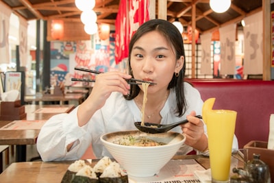 A person enjoys a bowl of ramen in a restaurant, holding chopsticks and a spoon. The interior is warmly lit with Japanese decor, including red lanterns and banners. A glass of orange juice and onigiri rice balls are also present on the table.