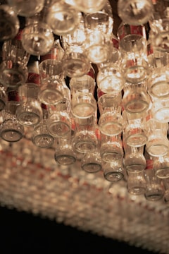 Dozens of glass bottles are arranged upside down, forming a chandelier-like structure. Light reflects and refracts through the bottles, creating a warm, ambient glow. The labels on the bottles are partially visible, hinting at their brand.