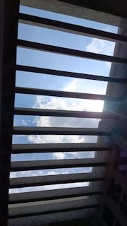 Concrete beams form a grid-like structure, framing a view of the sky filled with clouds. Sunlight partially penetrates the view, casting shadows and creating a play of light and dark across the structure.