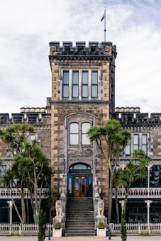 A grand stone castle-like building with a central tower and large windows, featuring detailed architectural elements. The entrance is flanked by stone statues on either side of a staircase. Tall palm trees are present in the foreground, adding a lush, tropical feel to the historic structure.