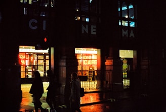 A nighttime street scene featuring the entrance to a cinema with illuminated signs. The building has a classic architectural style, and bright neon lights advertise a café inside. Silhouettes of people walking past are visible against the glow of the lights.
