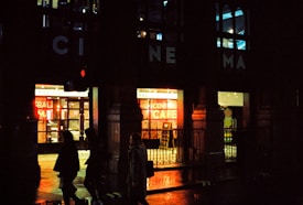 A nighttime street scene featuring the entrance to a cinema with illuminated signs. The building has a classic architectural style, and bright neon lights advertise a café inside. Silhouettes of people walking past are visible against the glow of the lights.