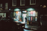 A dimly lit street scene featuring a newsagent shop with bright lights spilling from its window onto the wet pavement. A person stands inside near the entrance, while a black car is parked in front. Neon signs and posters are visible on the storefront.