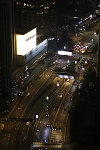 An aerial view of a busy urban highway at night with traffic flowing in both directions. On the left, a brightly lit building featuring signs such as 'The Intermark' and 'JP Morgan' stands out against the darkness. The roads are illuminated by streetlights and car headlights, with several vehicles visible on the road. The scene captures an intersection with traffic signals and city infrastructure visible in the background.
