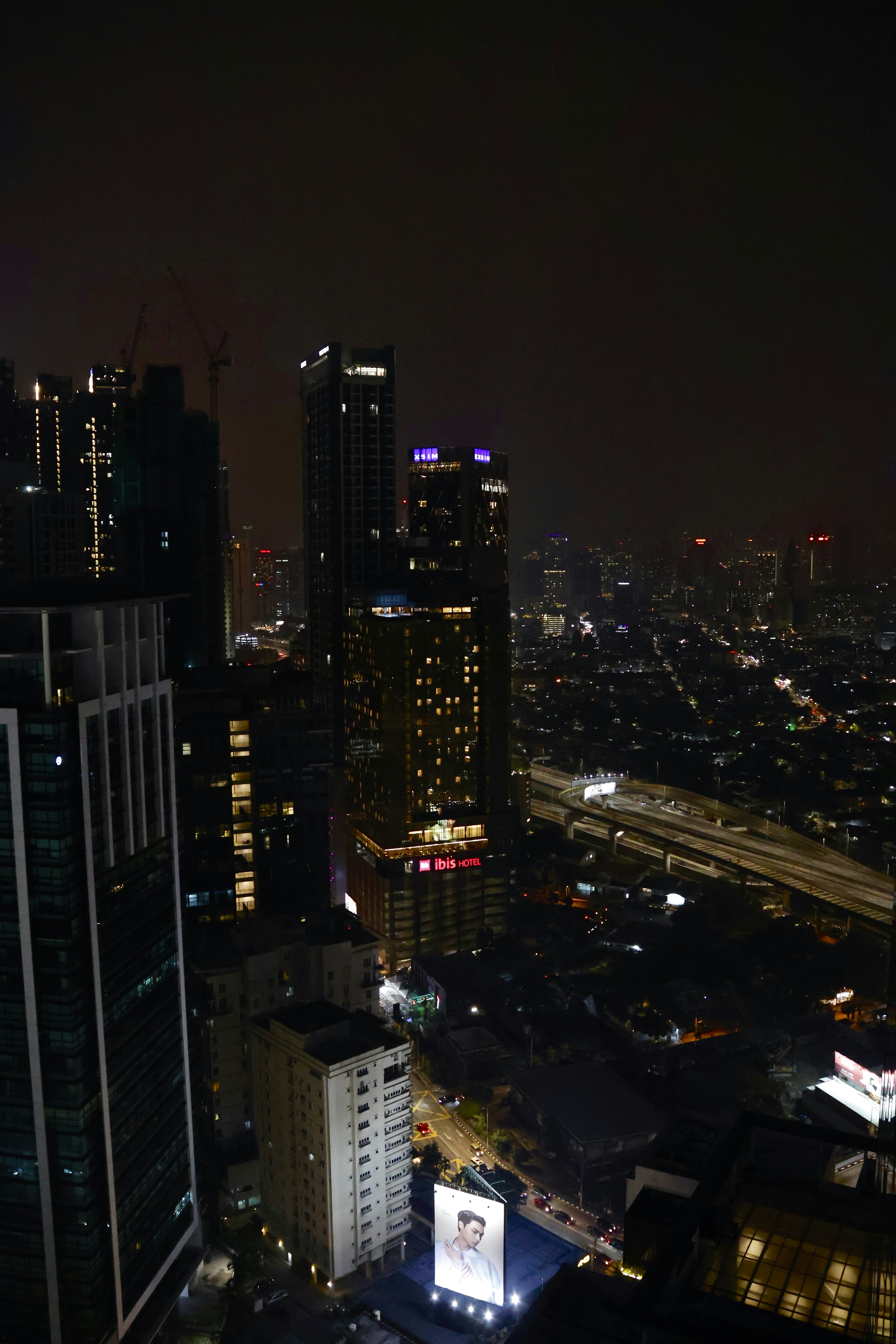 A nighttime cityscape featuring tall, illuminated skyscrapers. Neon signs and lights punctuate the dark skyline, with a large billboard displaying an advertisement at the bottom. The streets below show some levels of traffic, and the scene exudes urban energy.