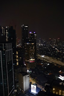 A nighttime cityscape featuring tall, illuminated skyscrapers. Neon signs and lights punctuate the dark skyline, with a large billboard displaying an advertisement at the bottom. The streets below show some levels of traffic, and the scene exudes urban energy.