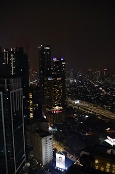 A nighttime cityscape featuring tall, illuminated skyscrapers. Neon signs and lights punctuate the dark skyline, with a large billboard displaying an advertisement at the bottom. The streets below show some levels of traffic, and the scene exudes urban energy.