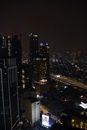 A nighttime cityscape featuring tall, illuminated skyscrapers. Neon signs and lights punctuate the dark skyline, with a large billboard displaying an advertisement at the bottom. The streets below show some levels of traffic, and the scene exudes urban energy.