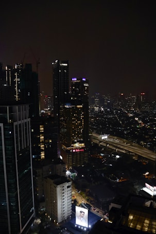 A nighttime cityscape featuring tall, illuminated skyscrapers. Neon signs and lights punctuate the dark skyline, with a large billboard displaying an advertisement at the bottom. The streets below show some levels of traffic, and the scene exudes urban energy.