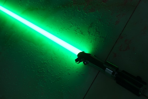 A brightly glowing green lightsaber rests on a textured surface, casting a vivid light. The hilt is metallic and detailed, contrasting with the vibrant blade.