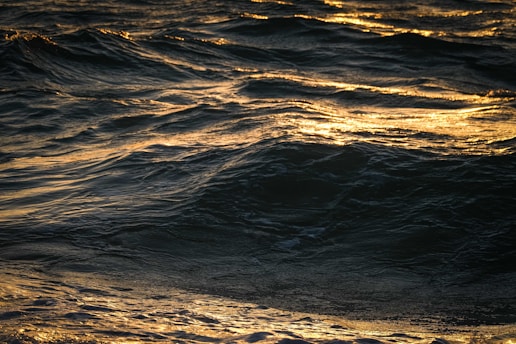 Waves of the ocean are illuminated by a warm golden light, creating a shimmering effect on the surface of the water. The crests and troughs of the waves are clearly visible, with some areas appearing darker where the light does not reach.