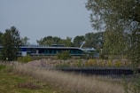 A modern building or structure with a sleek design is situated amidst a natural environment. It is surrounded by trees and vegetation, with a large expanse of grassy field in the foreground. A pile of stones or gravel is visible in front of the structure.