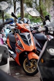 An orange scooter is parked among other scooters, featuring a sleek design with stylish graphics and gold-colored wheels. The background is filled with greenery and blurred outlines of more motorbikes.