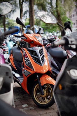 An orange scooter is parked among other scooters, featuring a sleek design with stylish graphics and gold-colored wheels. The background is filled with greenery and blurred outlines of more motorbikes.