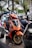 An orange scooter is parked among other scooters, featuring a sleek design with stylish graphics and gold-colored wheels. The background is filled with greenery and blurred outlines of more motorbikes.