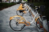 A row of rental bicycles with yellow and white designs parked on a paved pathway. The bikes are lined up in a docking station under trees, with grass on the sides of the path. Each bike features advertising and company branding on the rear wheel cover.