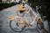 A row of rental bicycles with yellow and white designs parked on a paved pathway. The bikes are lined up in a docking station under trees, with grass on the sides of the path. Each bike features advertising and company branding on the rear wheel cover.