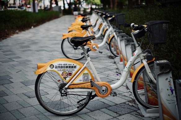A row of rental bicycles with yellow and white designs parked on a paved pathway. The bikes are lined up in a docking station under trees, with grass on the sides of the path. Each bike features advertising and company branding on the rear wheel cover.