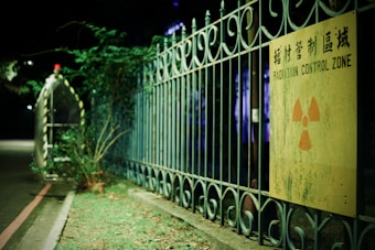 A dimly lit area featuring an iron fence with curly designs. Attached to the fence, a worn yellow sign displays a radiation symbol with the text 'Radiation Control Zone' in both English and Chinese. The ground is covered with some grass and there is a narrow road with a red line on the left.