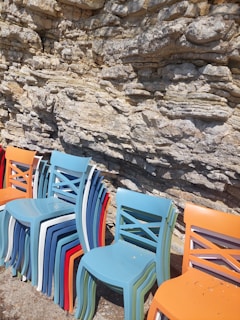 A stack of colorful plastic chairs is placed against a textured stone wall. The chairs are in various shades including blue, white, red, and orange. The stone wall features rough, layered patterns with irregular shapes and shadows.