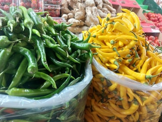 A vibrant display of fresh green and yellow chili peppers piled high in plastic bags. In the background, boxes of fresh produce such as tomatoes and ginger are visible.