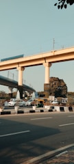 Elevated railway tracks supported by large pillars with a busy road below, featuring cars, a bright green and yellow auto-rickshaw, and trees. The sky is clear, and the setting appears urban with a combination of natural and man-made elements.