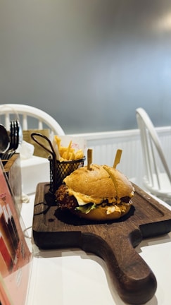 A delicious-looking burger with lettuce and sauce is placed on a rustic wooden serving board. Beside the burger, there is a small metal basket containing French fries. The setting appears to be indoors, with white furniture and soft, neutral lighting.