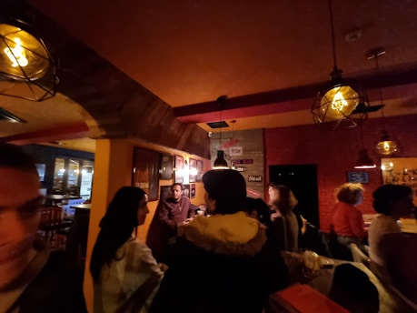 A dimly lit bar or restaurant with warm lighting and wooden decor, where people are socializing and sitting at tables and the bar. Several hanging light fixtures cast a soft glow over the space.