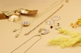 A collection of gold and silver jewelry is displayed on a light beige surface, including necklaces, rings, and earrings. A decorative gold bow and dried yellow flowers add a stylish touch to the arrangement, creating a luxurious and elegant aesthetic.