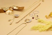 A collection of gold and silver jewelry is displayed on a light beige surface, including necklaces, rings, and earrings. A decorative gold bow and dried yellow flowers add a stylish touch to the arrangement, creating a luxurious and elegant aesthetic.