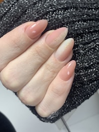 A hand with well-manicured nails painted in a natural, nude color with subtle glittery accents. The nails are shaped in an almond style, and the hand is resting on a black knitted fabric that provides a textured background.