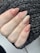 A hand with well-manicured nails painted in a natural, nude color with subtle glittery accents. The nails are shaped in an almond style, and the hand is resting on a black knitted fabric that provides a textured background.