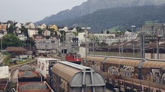 A railway yard with several cargo trains and a red locomotive on the tracks. The yard is busy with freight wagons filled with gravel and covered with graffiti. In the background, there are residential houses on a hillside, with a range of mountains further behind.