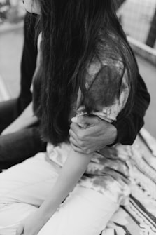 A black and white image featuring a close-up of two people sitting together. One person has their arm wrapped around the other, who has long hair and is wearing a patterned shirt. The background is slightly out of focus, giving the scene an intimate feel.