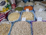 Various grains and legumes are displayed in open sacks, each labeled with a price tag. Items include chickpeas, lentils, beans, and other dry goods. There are also cleaning supplies and paper towels in the background.