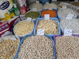 Various grains and legumes are displayed in open sacks, each labeled with a price tag. Items include chickpeas, lentils, beans, and other dry goods. There are also cleaning supplies and paper towels in the background.