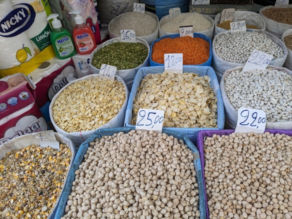 Various grains and legumes are displayed in open sacks, each labeled with a price tag. Items include chickpeas, lentils, beans, and other dry goods. There are also cleaning supplies and paper towels in the background.
