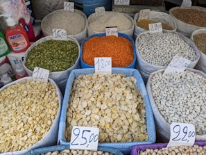 A variety of grains and legumes displayed in large sacks at a market, each labeled with a price tag. The sacks contain different types of beans, lentils, and seeds in various colors such as green, orange, white, and beige. The prices are written on cards placed on top of each sack. Nearby, there are bottles of detergent and paper towel rolls.