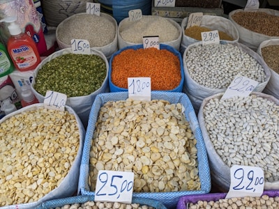 A variety of grains and legumes displayed in large sacks at a market, each labeled with a price tag. The sacks contain different types of beans, lentils, and seeds in various colors such as green, orange, white, and beige. The prices are written on cards placed on top of each sack. Nearby, there are bottles of detergent and paper towel rolls.